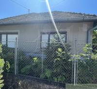 Rodinný dom v pôvodnom stave na 7 árovom pozemku v Tvrdošovciach - Rodinný dom v Tvrdošovciach s plotom a zeleňou, jasná slnečná obloha.