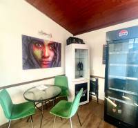 PRENÁJOM - Bistro/ Gastro prevádzka - Košice - Juh - Gastro priestory s podlahou s dreveným dekorom, zelené stoličky, obrazy a chladnička.