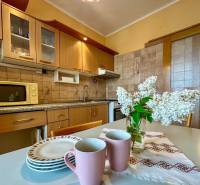 Predaj 3 - izbového bytu - Nová Baňa - Kuchyňa v 3-izbovom byte s drevenými skrinkami, kvetmi a riadom na stole.
