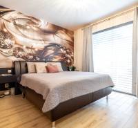NA PREDAJ SMART 4-IZB BUNGALOV SO ZÁHRADOU A VÝHĽADOM NA JAZERO - Spálňa s dekoratívnou stenou, veľkou posteľou a podlahou s dreveným dekorom v rodinnom dome.
