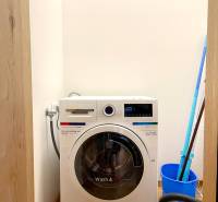 I 3 i byt s parkovaním v rezidenčnom komplexe METROPOLIS - Práčka so sušičkou v 3-izbovom byte, podlaha s dreveným dekorom, vedľa vedro a vysávač.