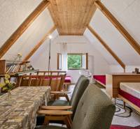 NA PREDAJ CHATA V REKREAČNEJ OBLASTI RUDAVA, OBEC MALÉ LEVÁRE - Podkrovie chaty s dreveným dekorom, stolom, pohovkou a posteľami pre útulnú atmosféru.