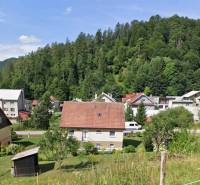 Veľkorysý 10-árový pozemok, slnečná nehnuteľnosť, priestranné bývanie  - Rodinný dom v Liptovských Revúcach obklopený zeleňou a ďalšími domami na pozadí lesa.