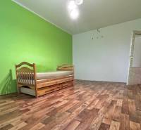 Na predaj 2i byt Levice - Izba s posteľou v 2-izbovom byte, podlaha s dreveným dekorom, zelené steny.