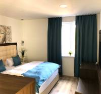 Na predaj apartmán s veľkou terasou Podhájská. - Garsónka so svetlým nábytkom, posteľou a modrými závesmi, podlaha s dreveným dekorom.