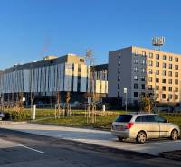 Bory:Na prenájom úplne nový, elegantný 2i byt/eng.text inside - Budovy na ulici Dominika v Lamači, Bratislava, obklopené parkoviskom a kopcami v pozadí.