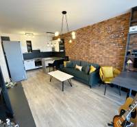 KOŠICE-2 IZBOVÝ BYT,NOVOSTAVBA,KOMPLET ZARIADENÝ,VL.PARK. MIESTO - Obývacia časť 2-izbového bytu s tehlovou stenou, podlahou s dreveným dekorom a kuchynským kútom.