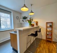 Tichý 2i byt v TOP Lokalite Nív | Trenčianska, BA - Kuchyňa v 2-izbovom byte s barovým pultom, podlahou s dreveným dekorom a závesnými lampami.