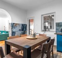 I Predaj štýlového tehlového domu v Rajke pri Bratislave - Jedáleň v rodinnom dome s dreveným stolom, modrou kuchynskou stenou a pohovkou v pozadí.