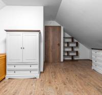 I Predaj štýlového tehlového domu v Rajke pri Bratislave - Biela skriňa, posteľ, zásuvky a poličky v podkroví rodinného domu s podlahou s dreveným dekorom.