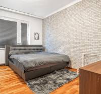 I Na predaj 2-i byt s terasou v Auersthal (Rakúsko) - Spálňa so sivou posteľou, tehlovou tapetou a podlahou s dreveným dekorom v 2-izbovom byte.