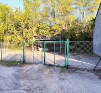 Prenájom oploteného pozemku – Nitra, Mlynárce – 390 m² - Plot s bránou a kovovou stavbou obklopený stromami v komerčnej časti Nitry.