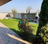 Apartmánový dom na predaj blízko termálneho kúpaliska - Na ulici neuvedené: záhrada s trávnikom, krb, obchodné priestory v pozadí.