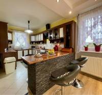 VKUSNÝ 4-IZBOVÝ RODINNÝ DOM S GARÁŽOU NA PREDAJ V OBCI TOPOĽNÍKY - Kuchyňa v rodinnom dome s barovým pultom, stoličkami a oknom s čipkovanými závesmi.