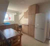 Apartmány na prenájom. - Moderná kuchyňa so svetlom z okna, drevenými skrinkami a stolom, biela budova.