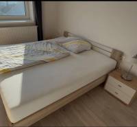 Prenájom 2-izbového bytu, Dunajská Streda - Manželská posteľ vedľa nočného stolíka s lampou, podlaha s dreveným dekorom.