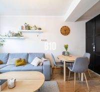 2-izbový byt po rekonštrukcii | Vietnamská ulica, Ružinov | Loggia | B - Obývačka so sivou sedačkou, okrúhlym stolom a podlahou s dreveným dekorom v 2-izbovom byte.