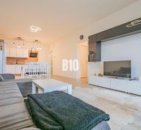 Výnimočný byt s panoramatickým výhľadom na Zobor - Obývacia izba s kuchyňou, biely nábytok, sedačka a podlaha s dreveným dekorom.