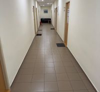 Ponúkame najlacnejšie  kancelárske priestory v Prievidzi  - Úzka chodba v kanceláriách s dlažbou, dverami a pohovkou na konci.