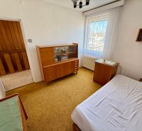 Rodinný dom s pivnicou, garážou a záhradou – Závod - Izba v rodinnom dome s drevenými skrinkami, posteľou, oknom s čipkovanými záclonami.