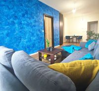 | Srdce Mesta | 2 Izb. byt | ORBIS |  70 m2 - Obývačka v 2-izbovom byte, sivý gauč, modrá stena, drevený dekor podlahy, jedálenský kút.