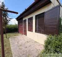 AREÁL 3 REKREAČNÝCH CHÁT PRI PODHÁJSKEJ – IDEÁLNE PRE DETSKÉ TÁBORY, R - Chata v Nových Zámkoch so zatvorenými drevenými okenicami a okolitou zeleňou na záhrade.