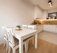 NOVOSTAVBA , BAŠTOVÁ 48 M2 2-IZBOVÝ BYT S LOGGIOU  - Kuchyňa v 2-izbovom byte s bielym stolom a podlahou s dreveným dekorom.