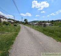 Slnečný stavebný pozemok 1 186 m² - Karná | možnosť dokúpiť 1 ha - Cesta obklopená trávou vedie cez vidiecku oblasť v Karnej s výhľadom na domy.