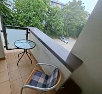 Priestranná garzónka s balkónom v novostavbe. - Balkón garsónky s výhľadom na zeleň a parkovisko, miestnosť so stolom a stoličkou.