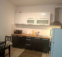 Priestranná garzónka s balkónom v novostavbe. - Kuchynská linka v garsónke so skriňami, jedálenský stôl, podlaha s dreveným dekorom.