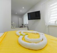 Moderný apartmán v centre Rajeckých Teplíc rovno oproti kúpeľom Afrodi - Svetlá izba Rekreačného apartmánu s televízorom, žltou posteľnou bielizňou a dekoráciou z uterákov.