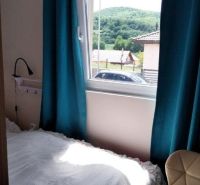 Mobilný celoročný drevodom – novostavba - Spálňa s jednoduchou posteľou, tyrkysovými závesmi a výhľadom na prírodu, Zvláštne nehnuteľnosti.