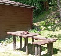 TURANY- rekreačná chata na pozemku 700m2 - Drevený stôl so stoličkou vedľa chaty, na ulici Černík. Lesná atmosféra, slnečný deň.