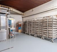 I PREDAJ – FUNGUJÚCA PEKÁREŇ A POTRAVINY V TRNAVE - Regály s kovovými vozíkmi a potravinovými zásobami vo výrobných priestoroch s podlahou z dreveným dekorom.