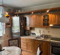 NA PREDAJ PRIESTRANNÝ 3-IZBOVÝ BYT | 67 m² | LODŽIA | 4. POSCHODIE - Kuchyňa v 3-izbovom byte s dreveným nábytkom, dekoratívnou záclonou a jedálenským stolom.