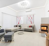 MODERNÁ NOVOSTAVBA | TOPOĽČANY | MRAVENISKO - Obývacia izba v rodinnom dome s krbom, sedačkou a kreslom, dekor červenými závesmi.