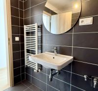 Na predaj novostavba 3 izbový byt, Bratislava, Nový Ružinov, Bajkalská - Kúpeľňa s tmavými dlaždicami, okrúhlym zrkadlom, umývadlom a rebríkovým radiátorom v 3-izbovom byte.