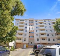 Prémiové bývanie v centre Nitry | 3 izby, veľký balkón - Bytový dom v Nitre obklopený stromami, viditeľné autá na parkovisku.