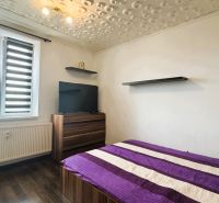 94,66 m², štyri izby a nikto nad hlavou. A Komenského má svoje výhody! - Spálňa v 4-izbovom byte s posteľou, televíziou, podlahou s dreveným dekorom a modernými žalúziami.