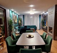 Znížená cena    Luxusný 4-izbový rodinný dom s letnou kuchyňou a nádhe - Jedálenský stôl so zelenými stoličkami, krb, pohovka a podlaha s dreveným dekorom.