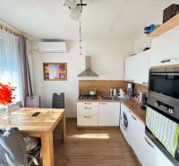 Prenajmem 1 izbový byt s balkónom v Nových Zámkoch - Kuchyňa v 1-izbovom byte s podlahou s dreveným dekorom, jedálenský stôl, okno s závesmi.