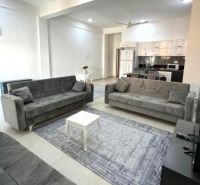 2-IZBOVÝ CONDO APARTMÁN PRI PLÁŽI, MBWENI, ZANZIBAR - Obývačka s tromi sivými pohovkami, stolíkom a kuchyňou v 2-izbovom byte.