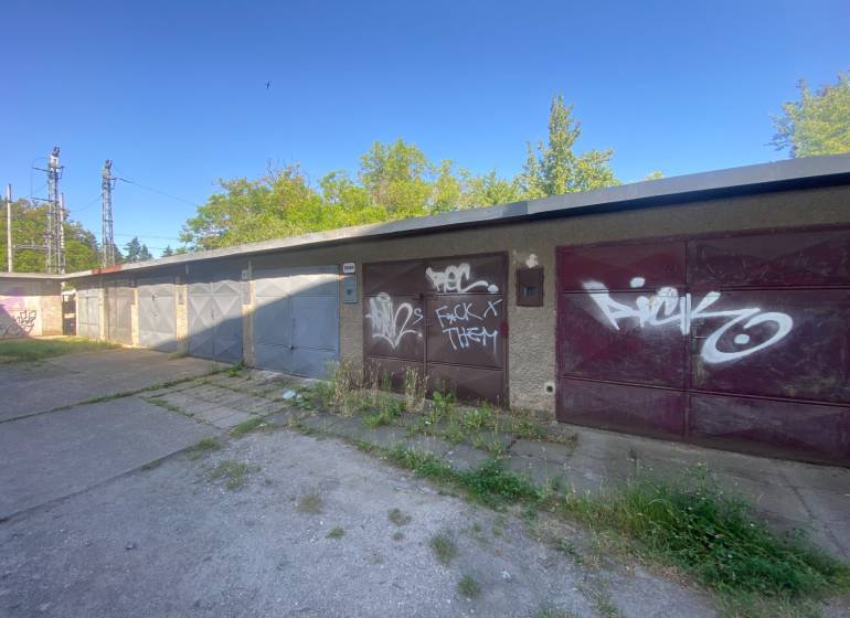 Garáž blízko Centra -  Zvolen na Predaj - Garáže vo Zvolene s grafitmi na bránach, obklopené zeleňou a betónovou plochou.