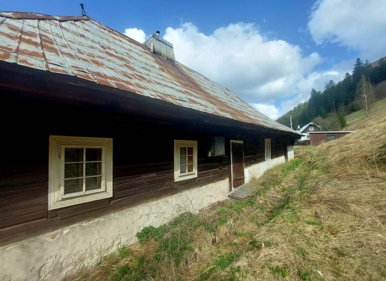 NA PREDAJ drevená chalúpka v Motyčkách  - Drevená chalupa s plechovou strechou na Motyčkách, obklopená prírodou a oblohou plnou oblakov.