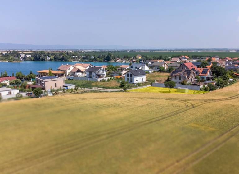  Stavebný pozemok pri jazere Nové Košariská  - Pozemky - bývanie v Dunajskej Lužnej s výhľadom na jazero, domy a pole.