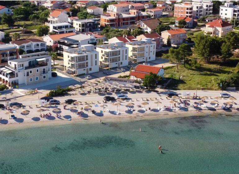CHORVÁTSKO - Luxusné apartmány (4i) - ostrov VIR - Pláž s lehátkami a slnečníkmi, za ňou domy a budovy v letovisku.