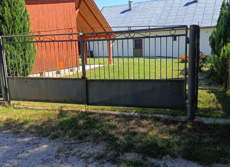 Chata s terasou a garážou TUŽINA okres Prievidza - Brána a drevená chata v Tužine, obklopené trávnikom a ihličnanmi.