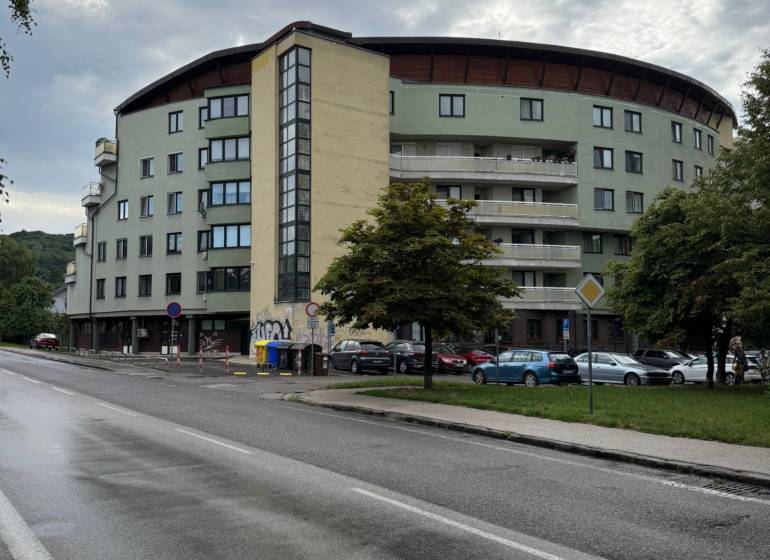 Garsónka v tehlovom dome. Dúbravka - Bytový dom v Bratislave - Dúbravke, obklopený ulicou, stromami a zaparkovanými autami.