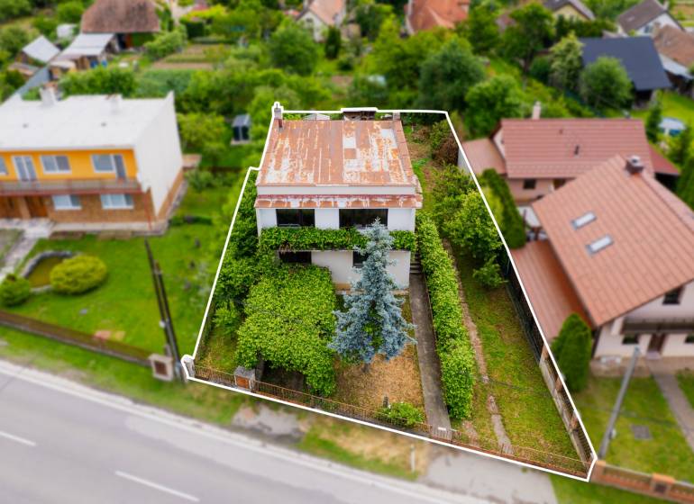 Na predaj Rodinný dom Drietoma – pozemok 745 m2  - Rodinný dom v Drietome obkolesený zeleňou a ďalšími domami, s plechovou strechou.