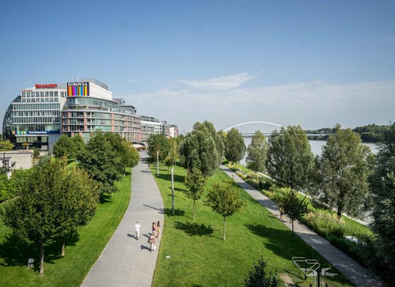 ,Predaj 3 izbový byt,100m2,Pribinova ulica,EUROVEA,Bratislava I - Eurovea v Bratislave, Nové Mesto, nábrežie rieky s chodníkom a zelenými stromami.